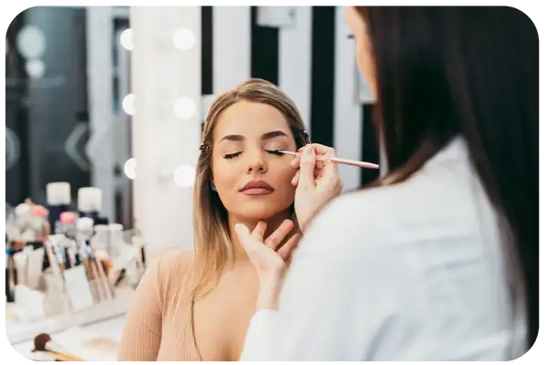 A woman with blonde hair is having her eyeliner applied by another person in a well-lit makeup station.